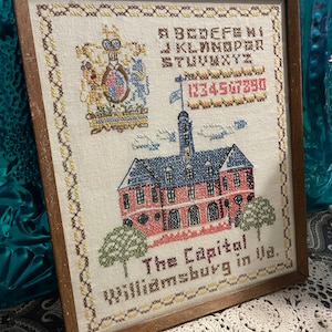 May include: A framed cross-stitch embroidery depicting the Capitol building in Williamsburg, Virginia. The embroidery includes a colorful coat of arms, an alphabet, and numbers. The text reads "The Capitol Williamsburg in Va."