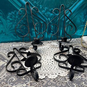 May include: Four black wrought iron candle holders. Two are wall-mounted with decorative scrollwork and a circular candle cup. The other two are tabletop candle holders with similar scrollwork and candle cups. The candle holders are on a patterned surface.