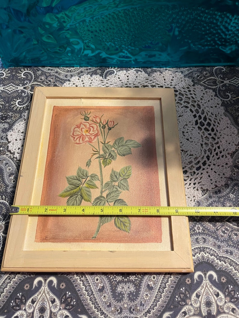 Puede incluir: Una pintura enmarcada de una rosa rosa con hojas y tallos verdes sobre un fondo marr&oacute;n claro.