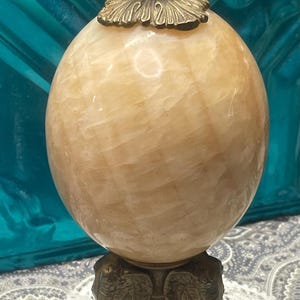 May include: A decorative sphere with a light brown, marbled appearance, topped with a bronze-colored finial. The sphere rests on a bronze stand with elephant-shaped legs, creating an elegant, vintage aesthetic.