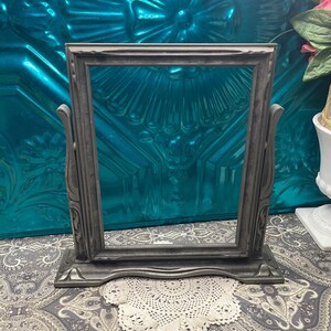 May include: A rectangular, dark gray metal picture frame with a swivel mechanism. The frame is supported by two curved side pillars attached to a wide, flat base. The frame features a smooth, matte finish with subtle decorative molding along the edges.