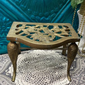 May include: A vintage brass trivet with a decorative floral and bird design. The trivet has four ornate legs and a rectangular top with cut-out details. The brass has a warm, aged patina, suggesting its antique nature.