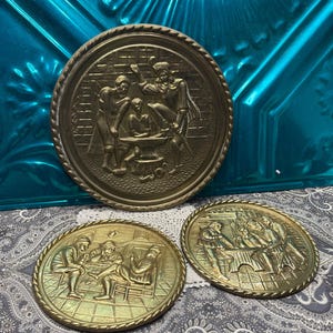 May include: Three round brass wall plaques featuring embossed scenes of historical figures. The largest plaque depicts a blacksmith at an anvil, while the two smaller plaques show groups of people gathered around tables in indoor settings.