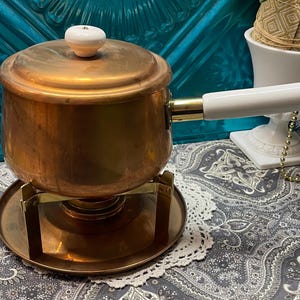 May include: A copper-colored fondue pot with a white handle and lid knob. The pot sits on a brass stand with a round base. The background includes a white decorative vase and a patterned fabric.