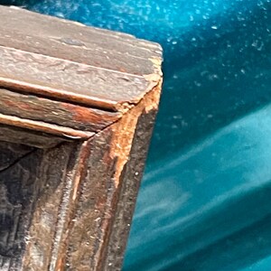 May include: Close-up of the corner of a wooden picture frame. The frame is dark brown with a reddish-brown patina. The wood is slightly weathered and shows signs of age.