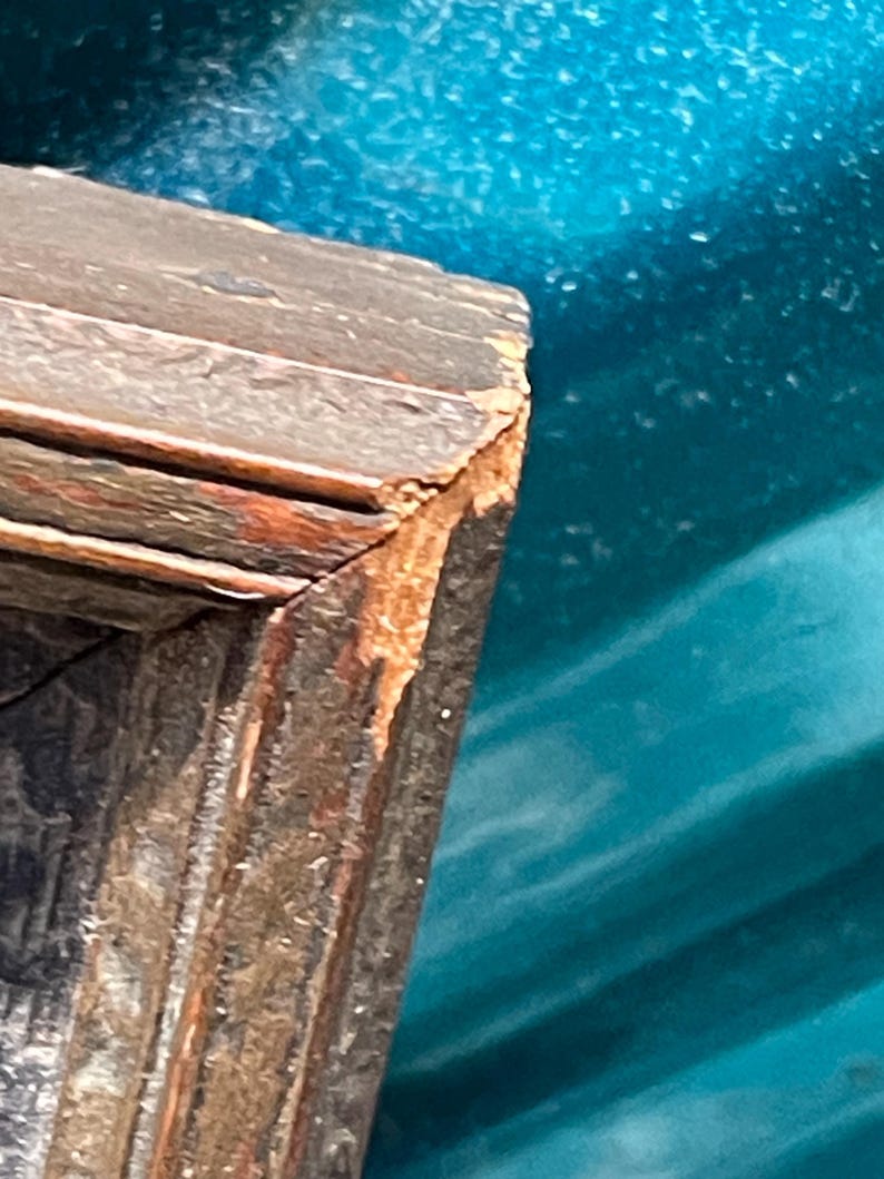 May include: Close-up of the corner of a wooden picture frame. The frame is dark brown with a reddish-brown patina. The wood is slightly weathered and shows signs of age.