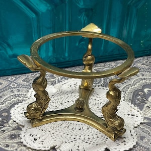 May include: A vintage brass sphere stand with a circular top and three dolphin-shaped supports. The stand has a detailed design and a warm, golden color, suitable for displaying decorative spheres or orbs.