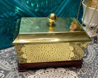 May include: A rectangular, gold-colored box with a matching lid and a round, gold knob. The box sits on a dark brown wooden base. The front of the box has a decorative, openwork design.