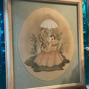 May include: A framed vintage watercolor painting of a couple standing under a white umbrella in a garden setting. The painting is in a light wood frame with a pale green mat. The couple is dressed in period clothing and the woman is holding a bouquet of flowers. The painting is signed "Mabel G. 1919".