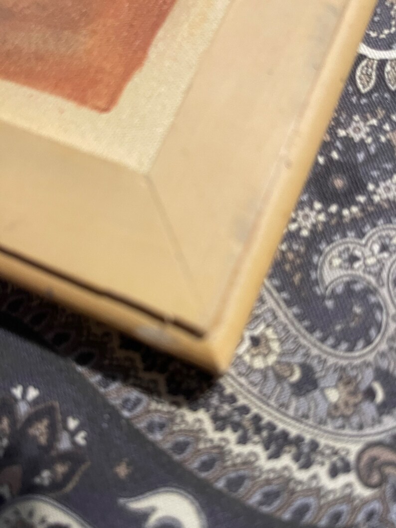 Puede incluir: Primer plano de la esquina de un marco de imagen beige. El marco es de madera y tiene un acabado liso. El marco est&aacute; sobre una tela estampada.