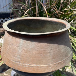 May include: A large, copper planter with a rounded bottom and a wide, flared rim. The planter has a rustic, aged appearance with a patina finish.