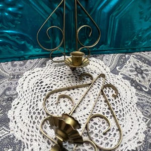 May include: Two vintage brass wall sconces with a scrolled design. Each sconce has a small cup for holding a candle. The sconces are displayed on a white lace doily and a patterned blue background.