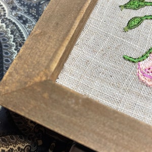 May include: A framed embroidery of pink flowers with green leaves on a beige linen background. The frame is made of brown wood.