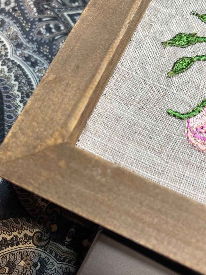May include: A framed embroidery of pink flowers with green leaves on a beige linen background. The frame is made of brown wood.