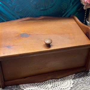 May include: A wooden bread box with a light brown finish. The box has a rectangular shape with a curved top and a small wooden knob. The sides have curved handles. The bread box is made of wood and has a natural wood grain.