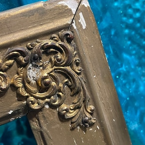 K&ouml;nnte beinhalten: Nahaufnahme eines goldbemalten Holzbildrahmens mit verzierten, goldfarbenen Metalldetails. Der Rahmen hat eine abgeplatzte Lackierung und zeigt Spuren des Alters.
