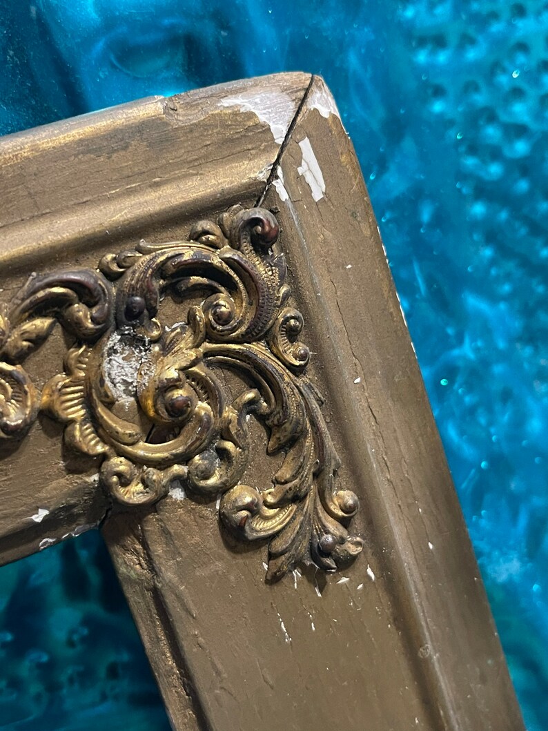 K&ouml;nnte beinhalten: Nahaufnahme eines goldbemalten Holzbildrahmens mit verzierten, goldfarbenen Metalldetails. Der Rahmen hat eine abgeplatzte Lackierung und zeigt Spuren des Alters.