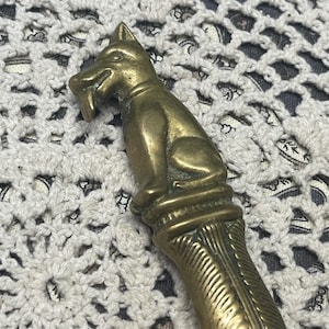 May include: A vintage brass letter opener featuring a seated dog figure at the top. The dog has a detailed face and is sitting atop a decorative column. The handle has a textured, ridged design.