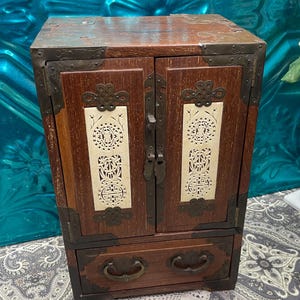 May include: A small, antique wooden cabinet with two doors and a drawer. The cabinet is a dark brown color with decorative metal corner brackets and hardware. The doors feature intricate, cream-colored cut-out designs. The drawer has two curved handles.