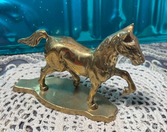 May include: A gold-colored metal horse figurine standing on a base. The horse is in a running pose with its head turned to the side.