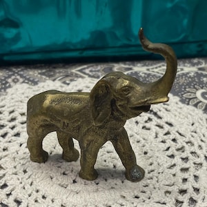 May include: A small, antique brass elephant figurine with its trunk raised. The elephant has detailed features, including large ears and a textured surface. The figurine is a decorative item, likely for display.