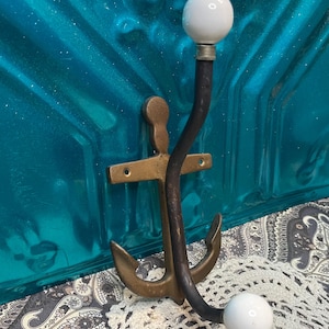 May include: A brass anchor-shaped coat hook with two white ceramic knobs. The hook is mounted on a turquoise wall.
