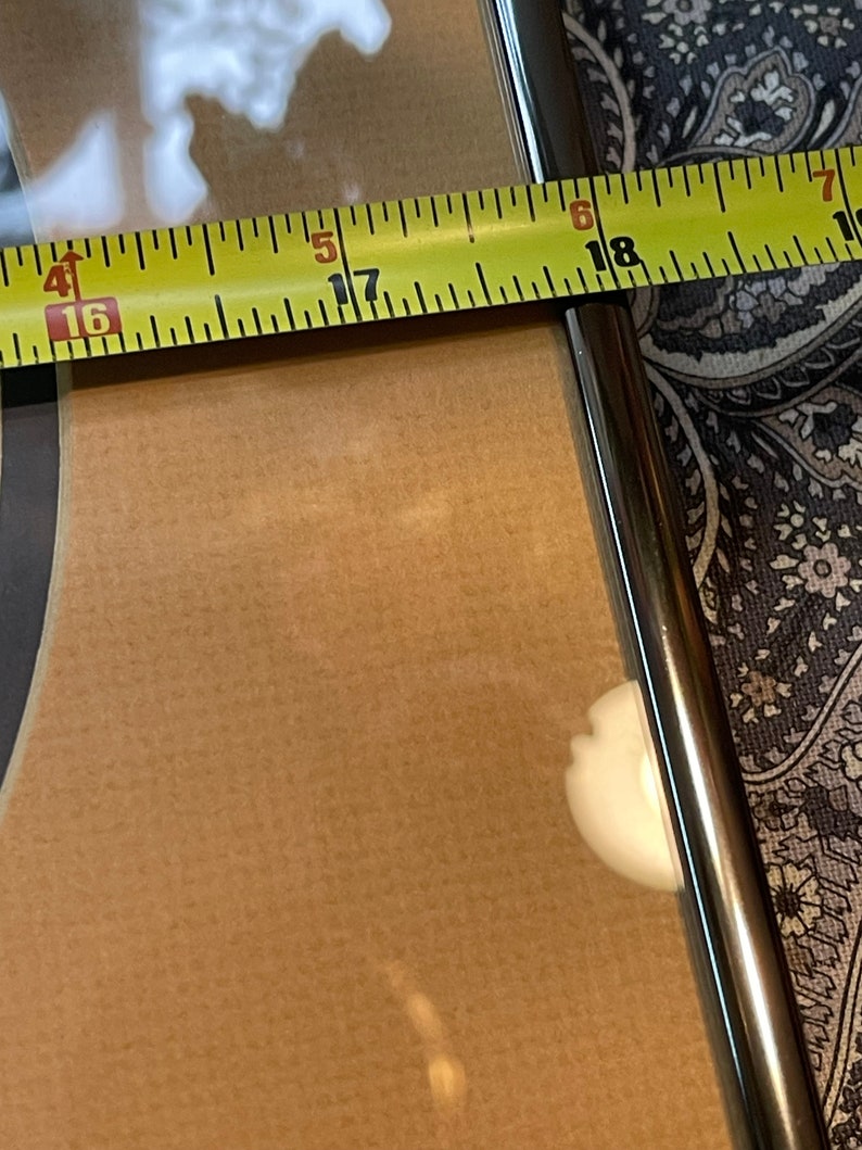 May include: A close-up of a yellow measuring tape with black markings. The tape is measuring the width of a brown wooden frame. The numbers 16, 17, 18 are visible on the tape.