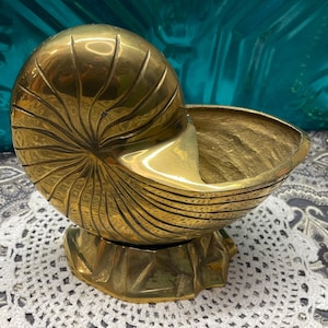 May include: A brass nautilus shell-shaped decorative bowl. The bowl has a spiral design and sits on a base. The bowl is a shiny gold color and appears to be made of metal. The bowl is on a white lace doily.