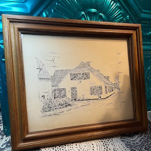 May include: A framed pen and ink drawing of a building with the word "Antik" written on the side. The artwork is in a brown wooden frame. The drawing depicts a street scene with a building and other architectural details.
