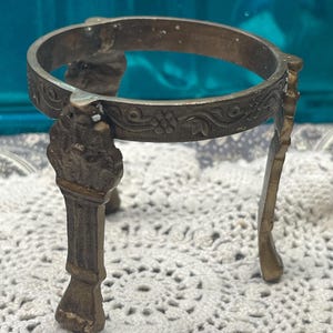May include: A circular bronze-toned metal display stand featuring three ornate legs and a decorative band with embossed floral and vine patterns. The stand has a weathered finish and is designed to hold spherical objects securely in place.