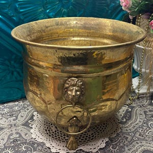 May include: A large, antique brass planter with a hammered texture. The planter features a lion's head detail with a ring handle and a decorative base. The brass has a warm, golden hue, and the planter sits on a white lace doily.
