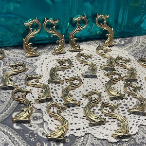 May include: A collection of 20 gold-toned metal decorative hooks with a swirling, ornate design. The hooks are arranged on a white lace doily with a paisley pattern.