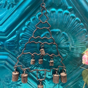 May include: A rustic metal Christmas tree ornament with small, aged bells hanging from its branches. The tree is suspended by a chain against a textured teal background, creating a vintage aesthetic. The bells are cylindrical and have a weathered appearance.