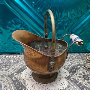 May include: A vintage copper coal scuttle with a brass handle and a decorative porcelain handle. The scuttle has a round base and a detailed design on the front. The interior shows signs of age and use.