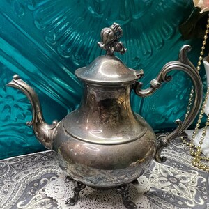May include: A silver-colored teapot with ornate detailing, including a decorative handle and a lid with a floral design. The teapot sits on a three-legged base and is displayed on a patterned surface.