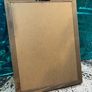 May include: The back of a brown wooden picture frame with a brown cardboard backing. The frame has a metal hanging hook on the top.