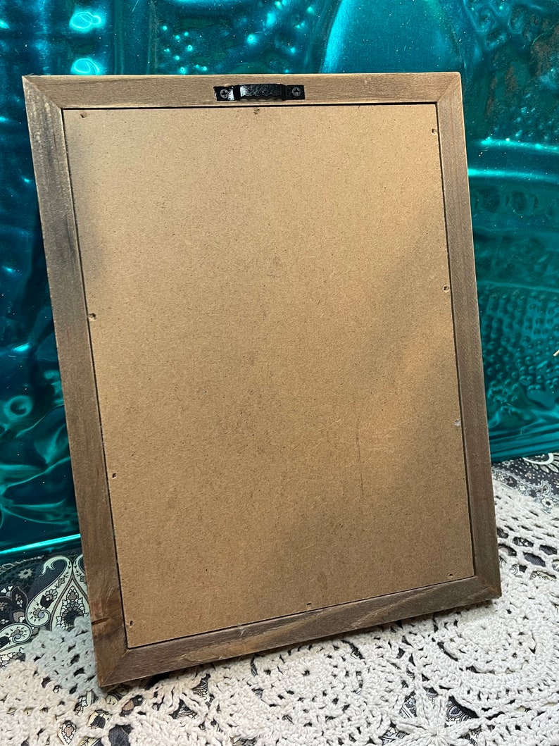 May include: The back of a brown wooden picture frame with a brown cardboard backing. The frame has a metal hanging hook on the top.