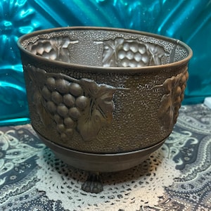 May include: A vintage metal planter with a grapevine design. The planter is brown and has a textured surface. It has a round base and three legs.