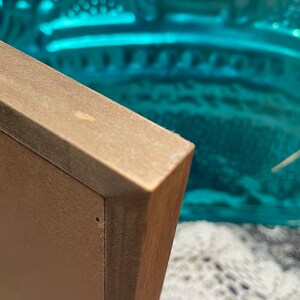 May include: Close-up of the corner of a brown wooden picture frame. The frame is made of wood with a light brown finish. The frame is in focus, while the background is blurred.