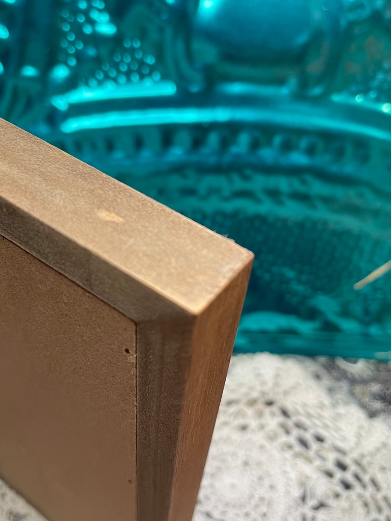 May include: Close-up of the corner of a brown wooden picture frame. The frame is made of wood with a light brown finish. The frame is in focus, while the background is blurred.