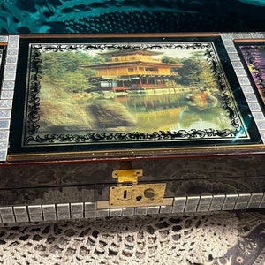 May include: A wooden box with a hinged lid and a lock. The box is decorated with three images: a Japanese garden, a woman in a traditional kimono, and a woman in a red dress. The box is made of dark wood and has a silver trim.