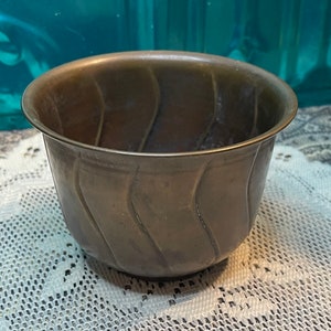 May include: A small, round, brass bowl with a wavy, textured design. The bowl is a dark brown color and has a shiny finish.