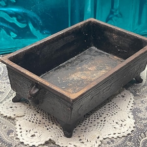 May include: A rectangular, dark brown cast iron vessel with a textured exterior, a smooth interior, and four small feet. The rim features a slightly lighter brown tone, and the sides are decorated with subtle, raised relief patterns.