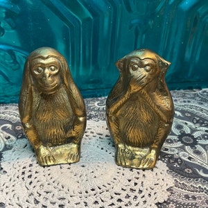 May include: Two gold-colored monkey figurines. One monkey is covering its eyes and the other is covering its ears. The monkeys are sitting on a white crocheted doily.