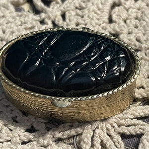 May include: A small, oval-shaped, black, carved, decorative box with a gold metal rim and intricate floral designs.