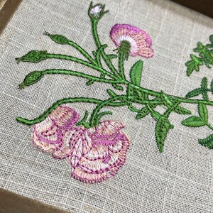 May include: Embroidered floral design on a beige linen fabric, featuring pink flowers and green leaves. The design is framed in a brown wooden frame.