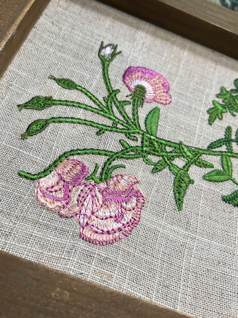 May include: Embroidered floral design on a beige linen fabric, featuring pink flowers and green leaves. The design is framed in a brown wooden frame.