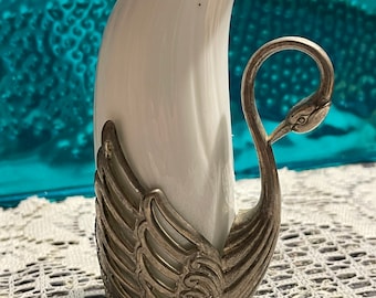 May include: A white ceramic vase shaped like a tooth is held by a silver swan figurine. The swan has intricate details and a delicate design.