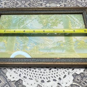 May include: A vintage framed print with a light blue and green landscape scene. The frame is made of wood with a dark brown finish and a gold trim.