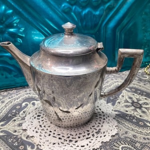 May include: A silver teapot with a lid and a handle. The teapot has a spout and is sitting on a white doily. The teapot has a decorative pattern on the side.